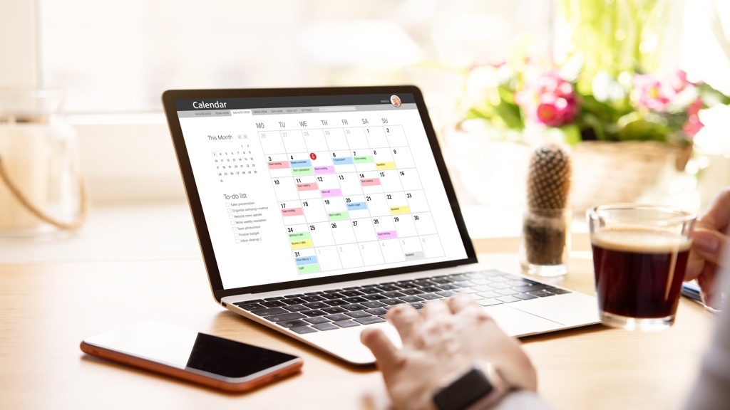 En person använder en bärbar dator som visar en digital kalender medan de håller en kopp kaffe i ena handen.