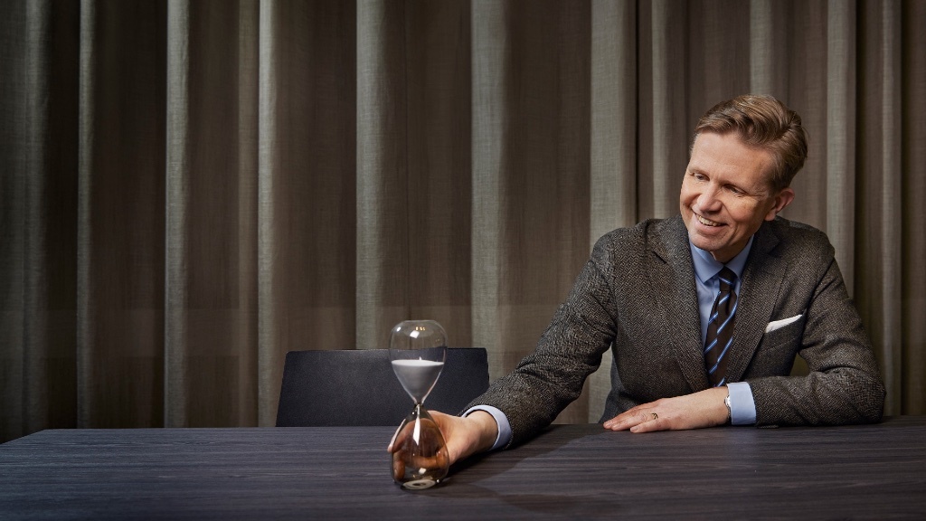 A smiling man in a suit sits at a table, gently holding an hourglass with one hand.