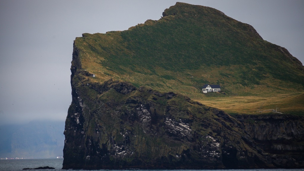 Ett litet vitt hus ligger ensligt på en gräsbevuxen, brant ö omgiven av klippor och havet.