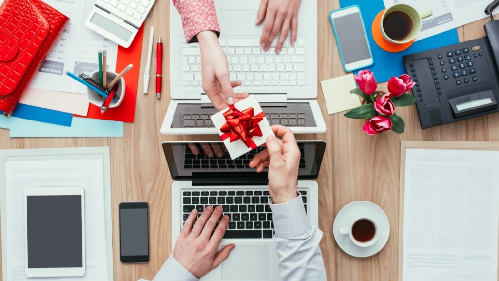 Two people exchange a small gift with a red bow over a desk filled with office supplies, laptops, and coffee cups.