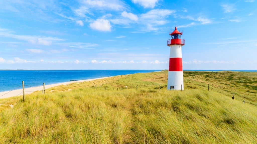 Ett rödvitt randigt fyrtorn står på en gräsbevuxen sanddyn med utsikt över havet under en klarblå himmel.