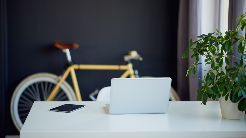 En minimalistisk arbetsyta med en laptop och en surfplatta på ett vitt skrivbord, bredvid en grön växt, med en cykel i bakgrunden.