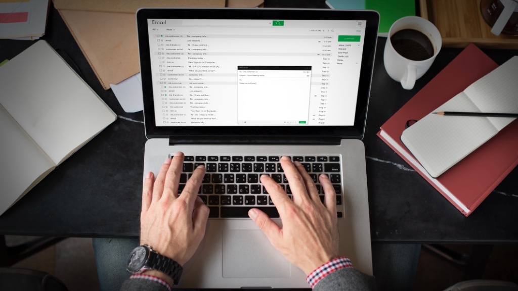 En person skriver på en laptop med ett e-postgränssnitt öppet, omgiven av en anteckningsbok, en kaffekopp och kontorsmaterial.