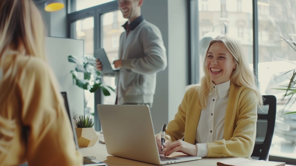 En blond kvinna ler medan hon sitter vid sitt skrivbord i en modern kontorsmiljö medan en manlig kollega närmar sig henne med en surfplatta i handen.