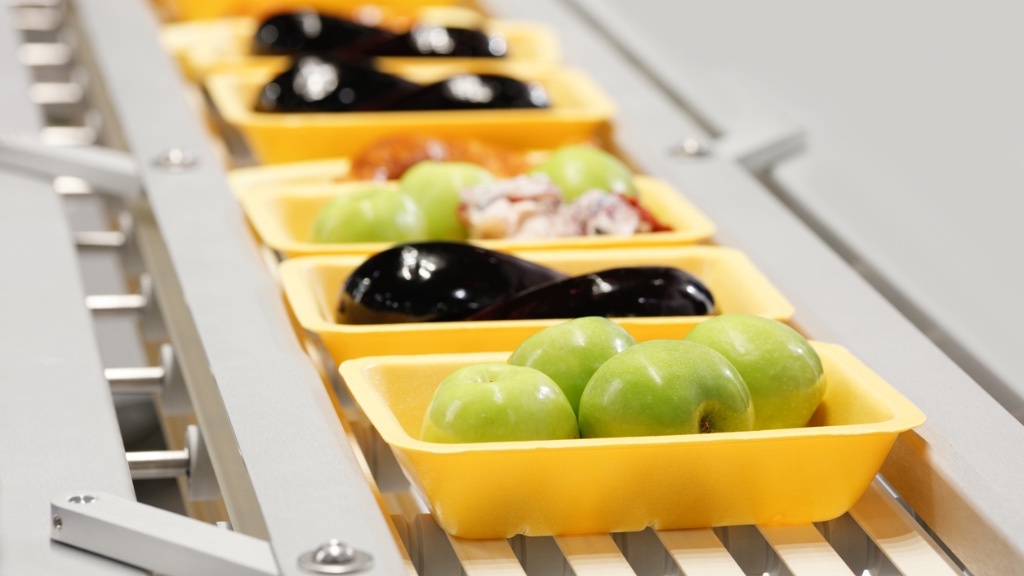 A conveyor belt carries yellow trays filled with green apples, eggplants, and other food items in a food processing or packaging facility.