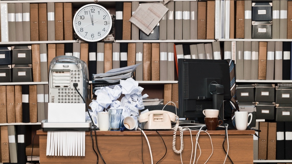 A cluttered office desk is surrounded by stacks of disorganized papers, books, mugs, and outdated office equipment, with an overloaded shelf in the background.