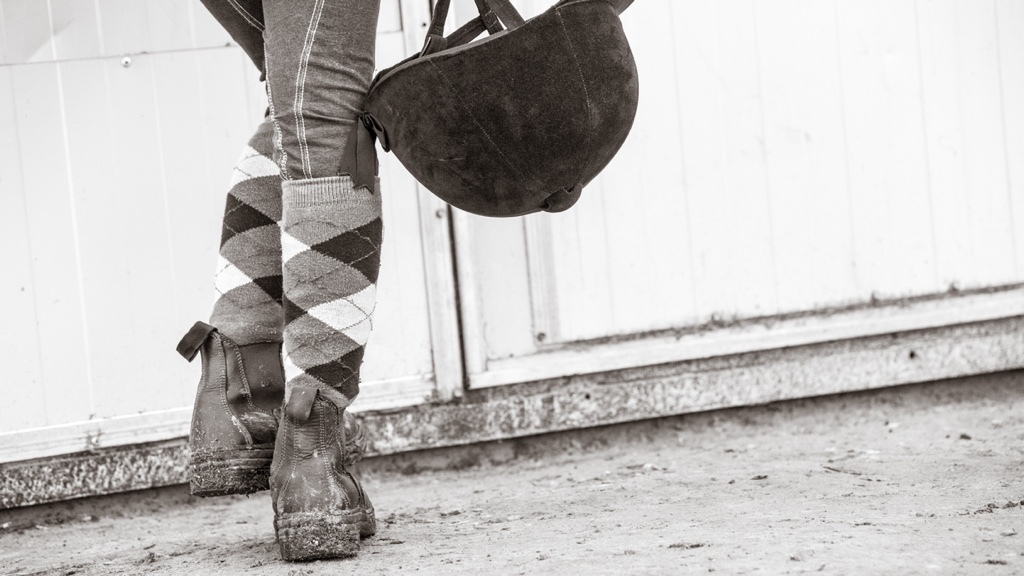 En person i mönstrade strumpor och leriga stövlar håller en ridhjälm vid sin sida nära en vit stallvägg.