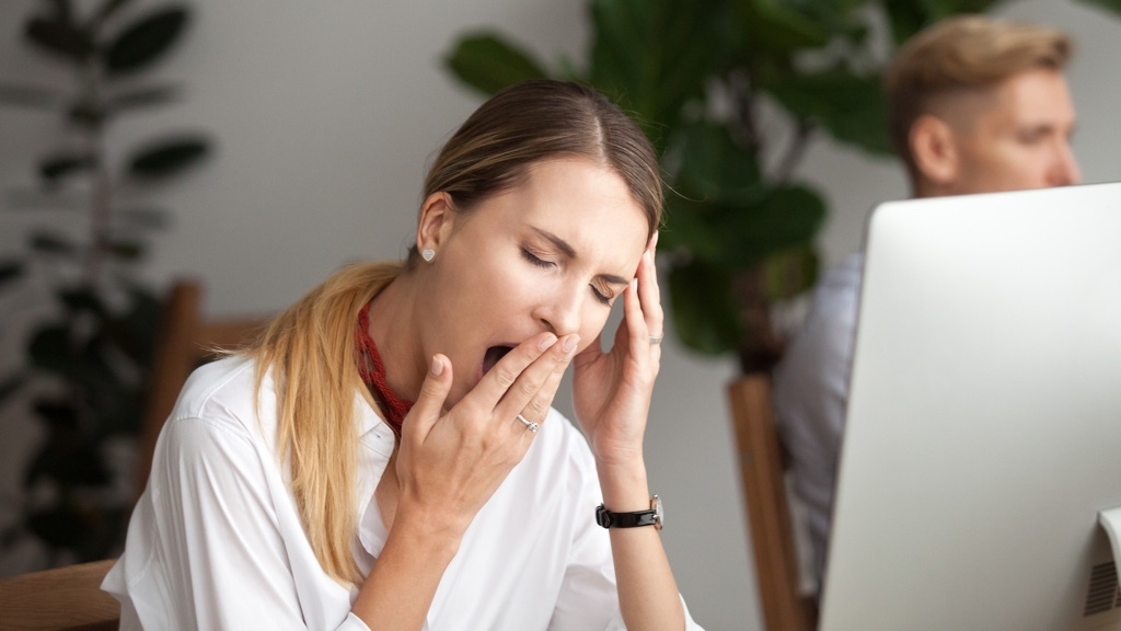 A woman is sitting at her desk yawning with her hand on her head, while a man works at another desk in the background.