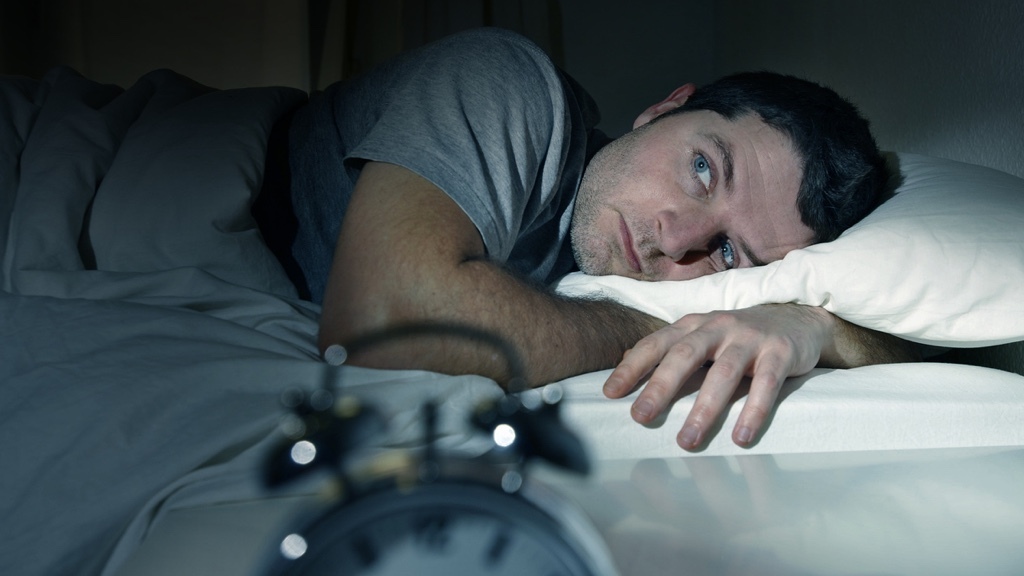 A man lies awake in bed, staring at an alarm clock with a look of sleeplessness.