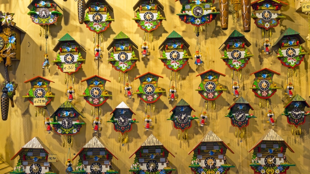 A colorful wall display of traditional Swiss-style cuckoo clocks, each adorned with intricate details and small figurines, is organized neatly, with prices visible on some of them.