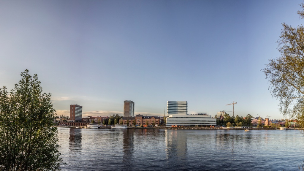 En naturskön vy över Umeås silhuett längs floden, med moderna byggnader och en klarblå himmel.