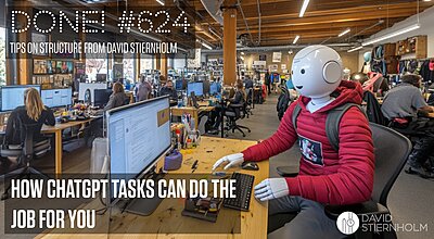 A humanoid robot wearing a red hoodie is sitting at a desk in a busy open-plan office, working on a computer.