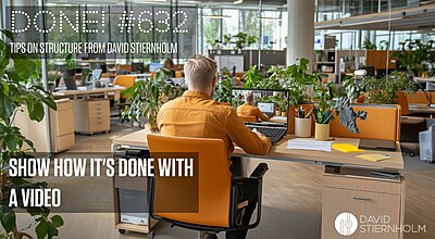 A man wearing an orange shirt sits at a modern office desk with an orange chair, working on his laptop, surrounded by green plants in an open-plan workspace.